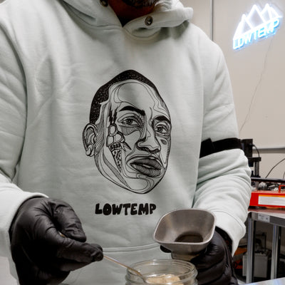 Person wearing a hoodie with a face design and 'LOWTEMP' text, holding a small container.