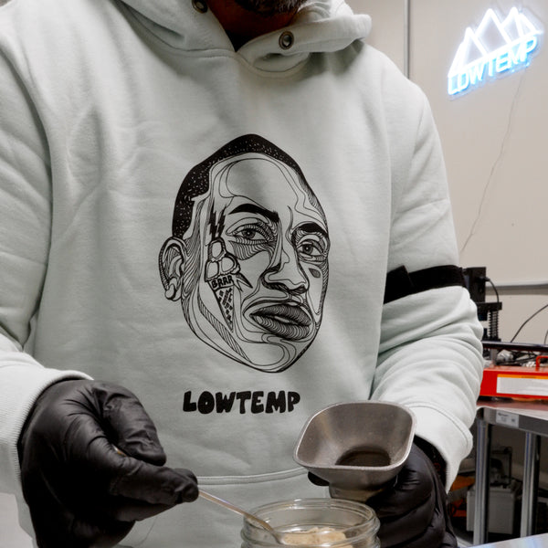 Person wearing a hoodie with a face design and 'LOWTEMP' text, holding a small container.