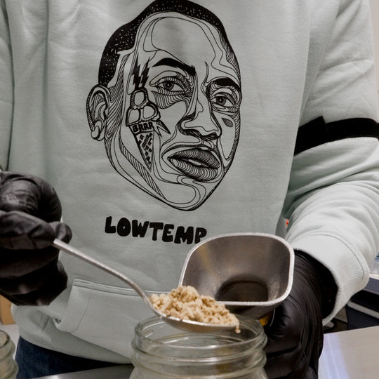 Person wearing a hoodie with a face design and 'LOWTEMP' text, holding metal spoons over a jar.