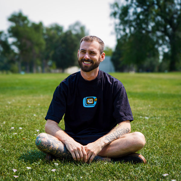 Man sitting on grass wearing a black t-shirt with a logo, smiling outdoors.